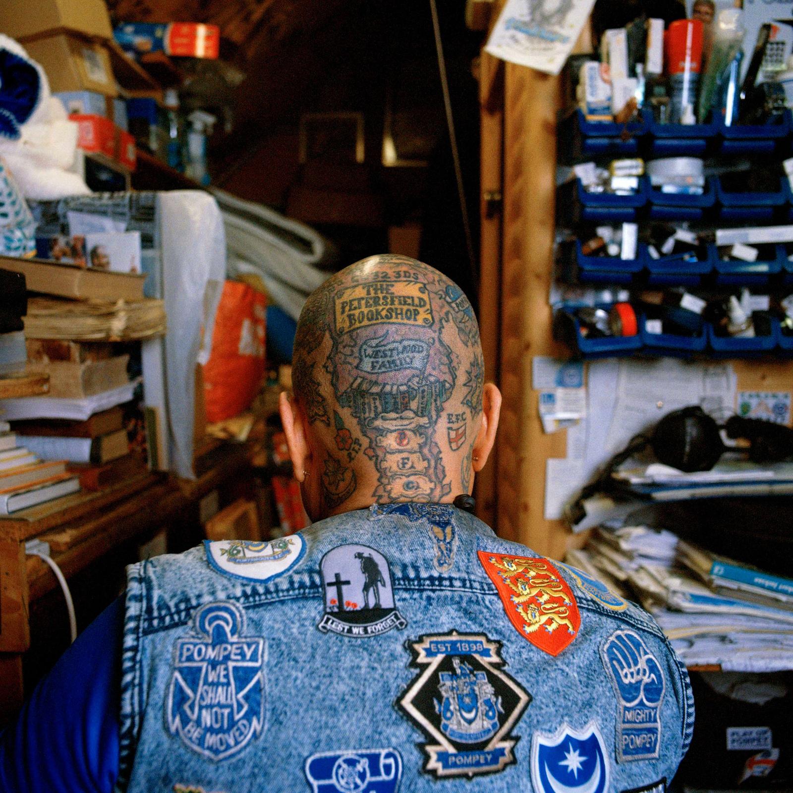 A person with tattoos stands in a cluttered room with books and boxes, wearing a denim jacket with patches and a leather jacket.