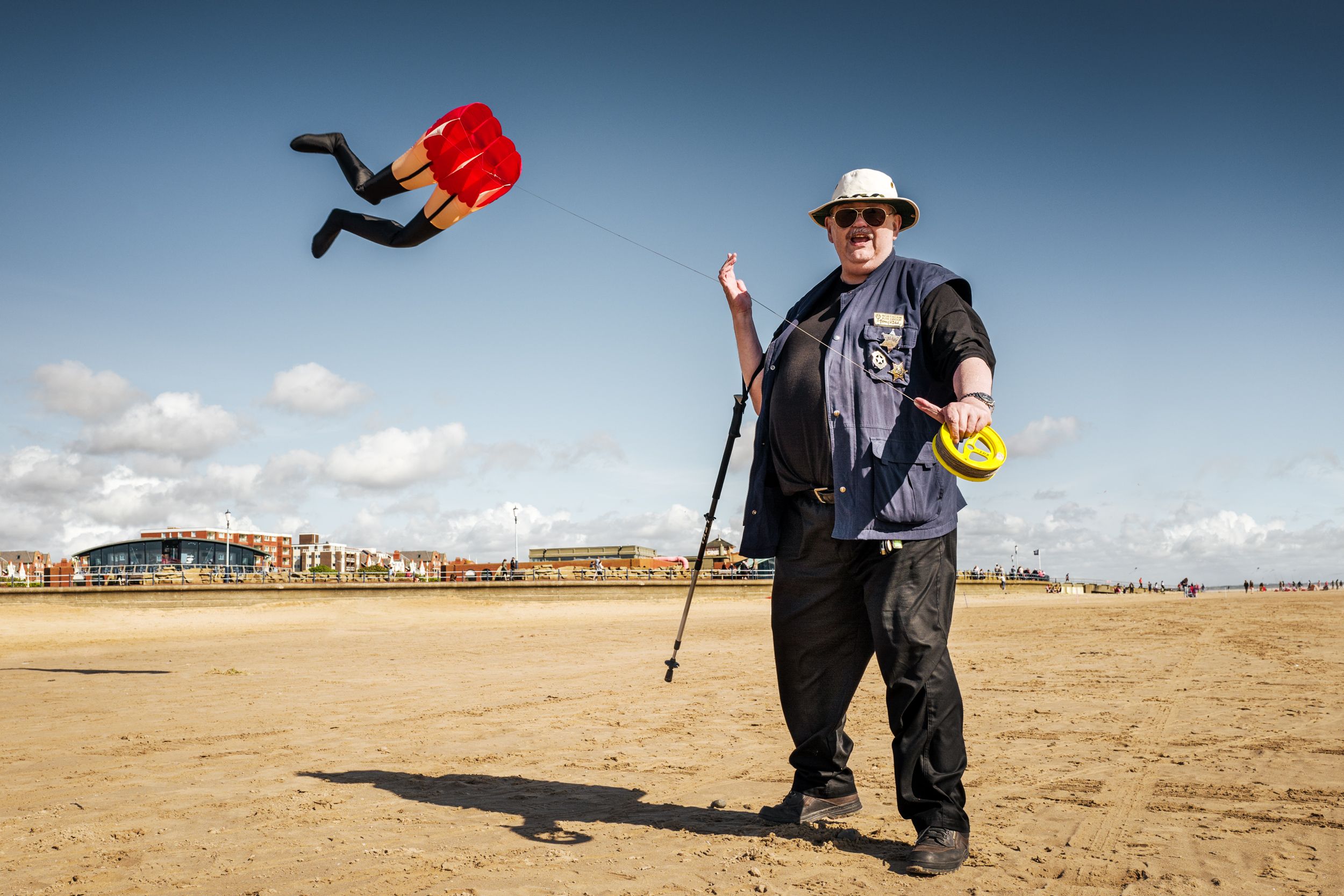 A man in a blue vest and hat is flying a red and yellow kite on a beach.