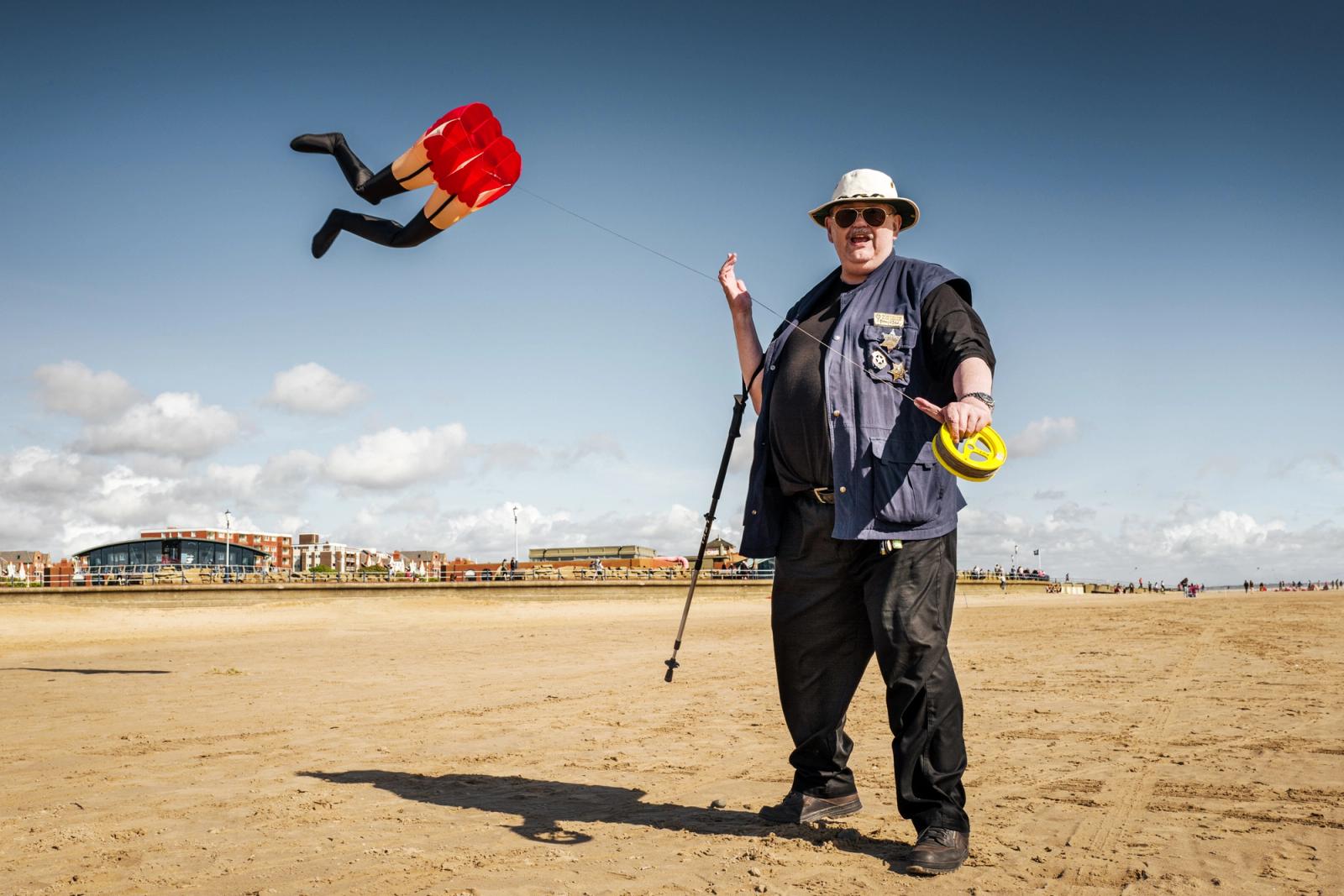 A man in a blue vest and hat is flying a red and yellow kite on a beach.