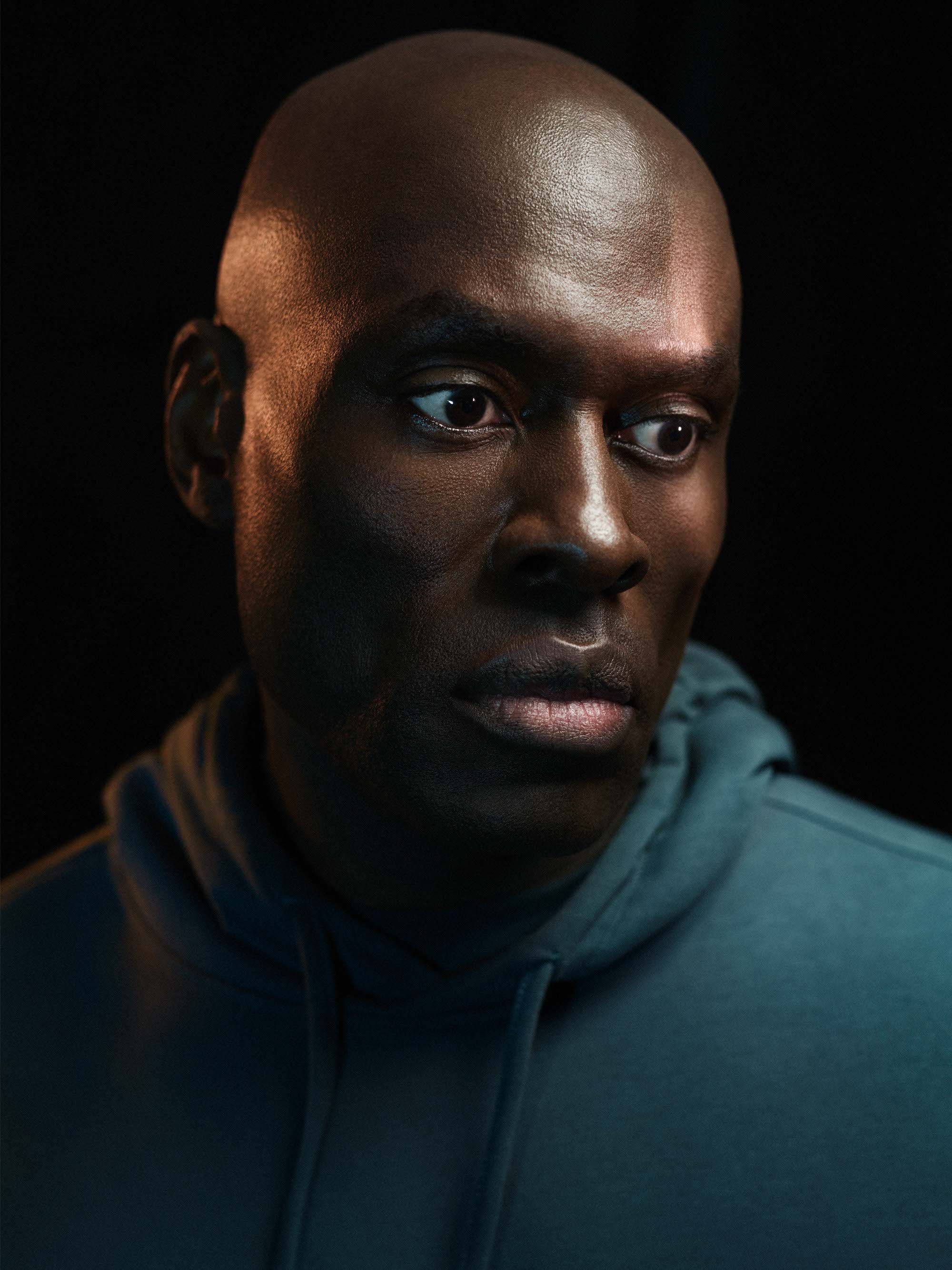 A close-up portrait of a bald, dark-skinned man with a serious expression, wearing a blue hoodie.