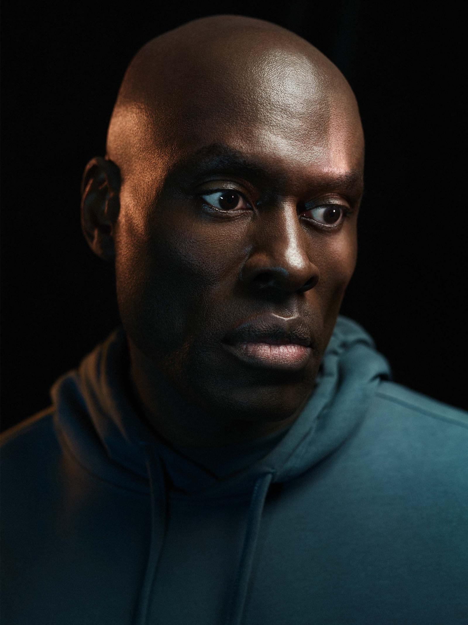 A close-up portrait of a bald, dark-skinned man with a serious expression, wearing a blue hoodie.