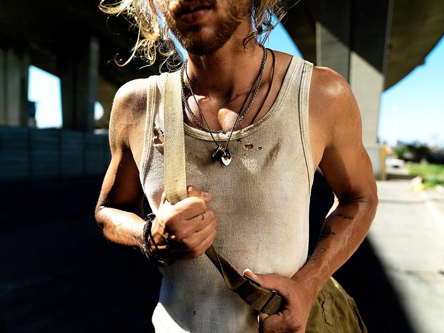 A bearded man in a white tank top stands in a dimly lit industrial space with concrete and metal backgrounds.