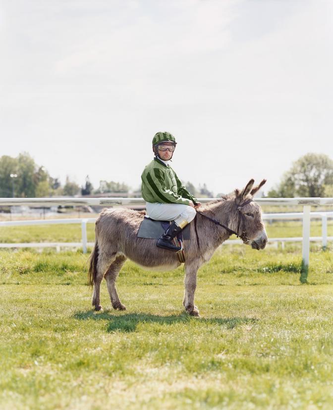 A person wearing a green and white striped shirt, riding a donkey in a green grassy field.