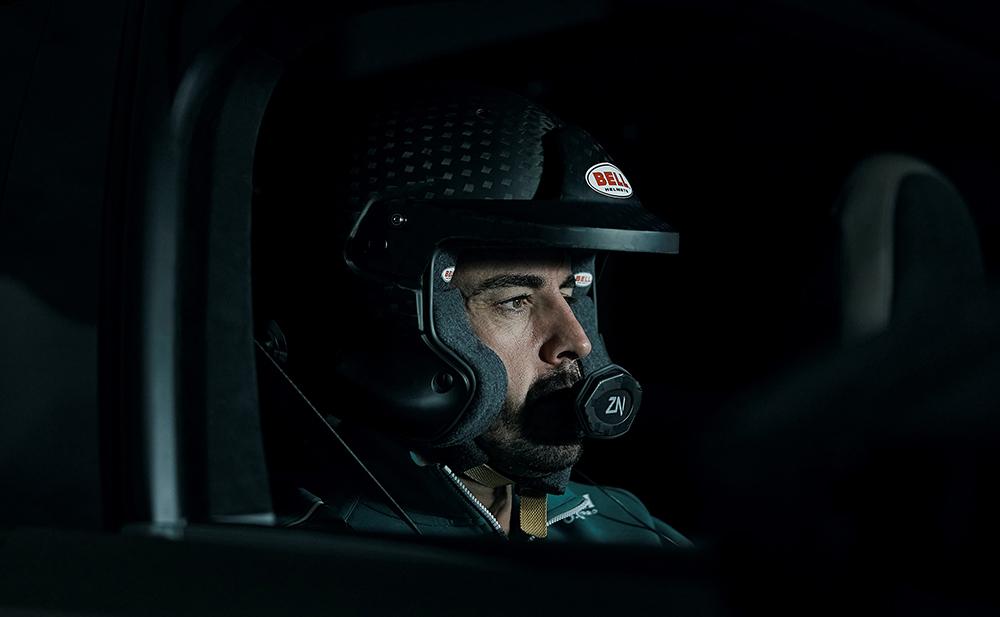 A man wearing a racing helmet and a black and white racing suit is sitting in a racing car, looking focused and ready to start the race.