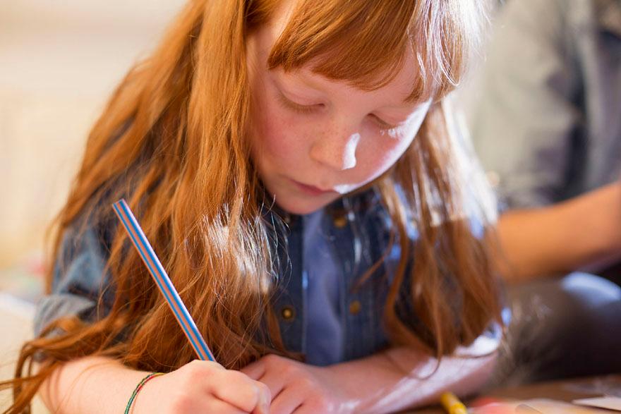 A young girl with long red hair is drawing with a blue pencil.