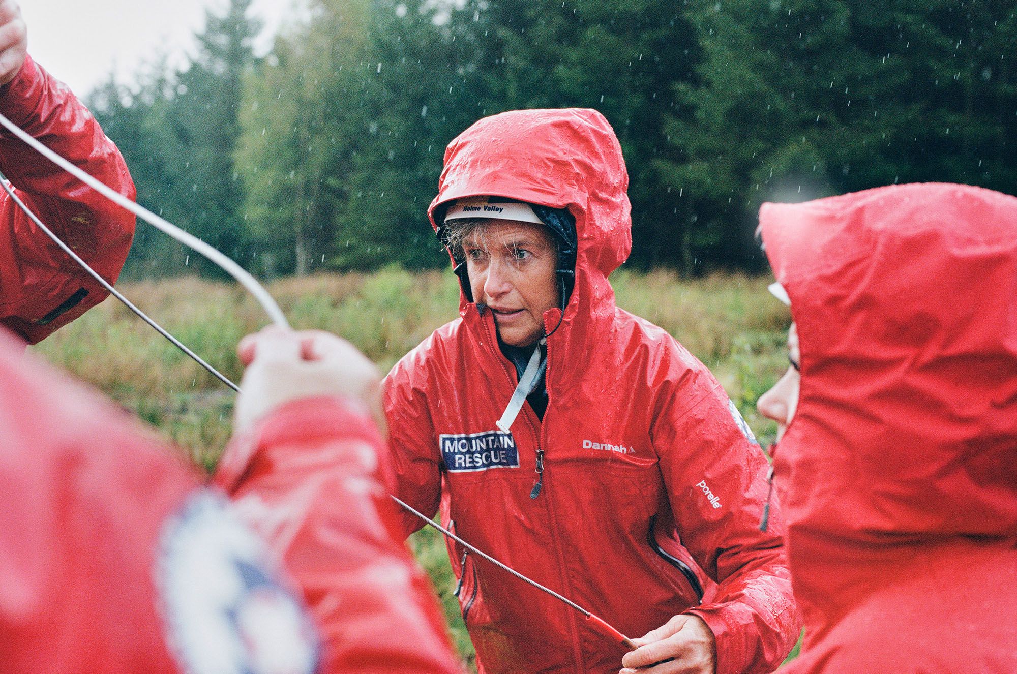 A group of people in red rain jackets and helmets are standing in a field, holding ropes and looking up at something in the distance.