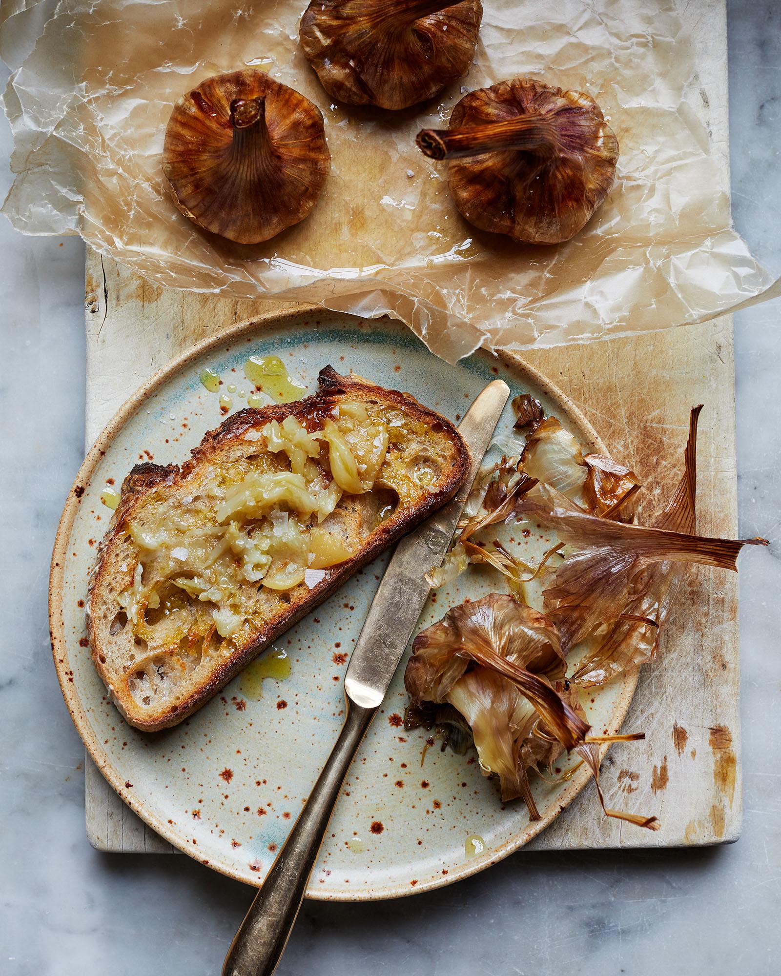 A slice of bread topped with roasted garlic and a knife on a plate, with a few roasted garlic bulbs in the background.