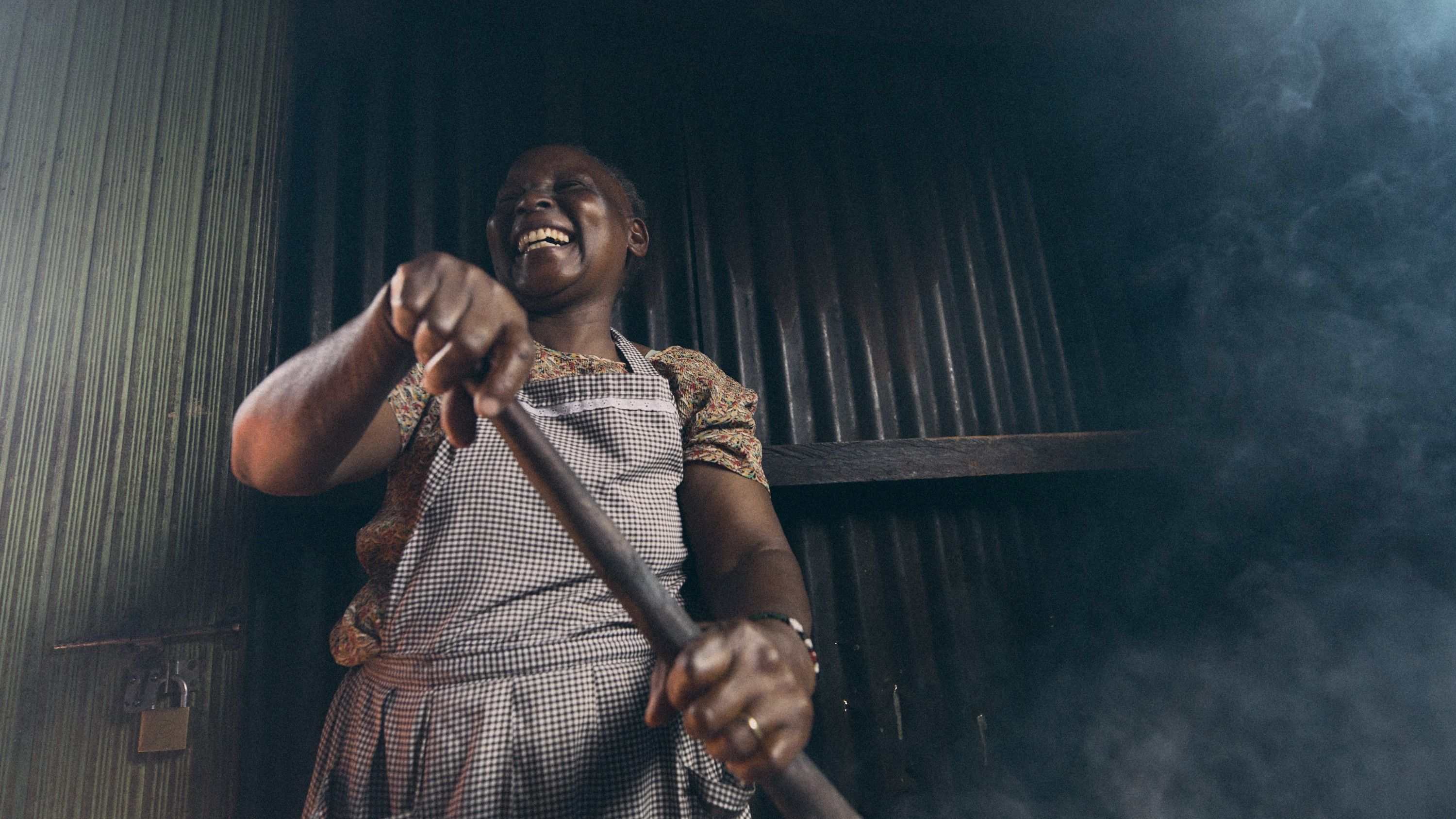 A woman in a white and black checkered apron, likely a chef or cook, is smiling and holding a long wooden spoon in a dimly lit, possibly industrial or commercial kitchen setting.