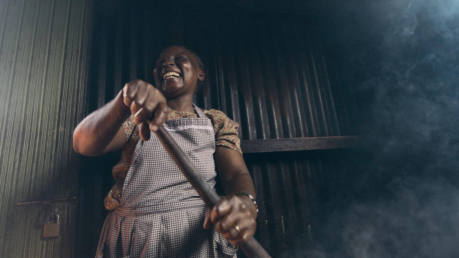 A woman in a white and black checkered apron, likely a chef or cook, is smiling and holding a long wooden spoon in a dimly lit, possibly industrial or commercial kitchen setting.