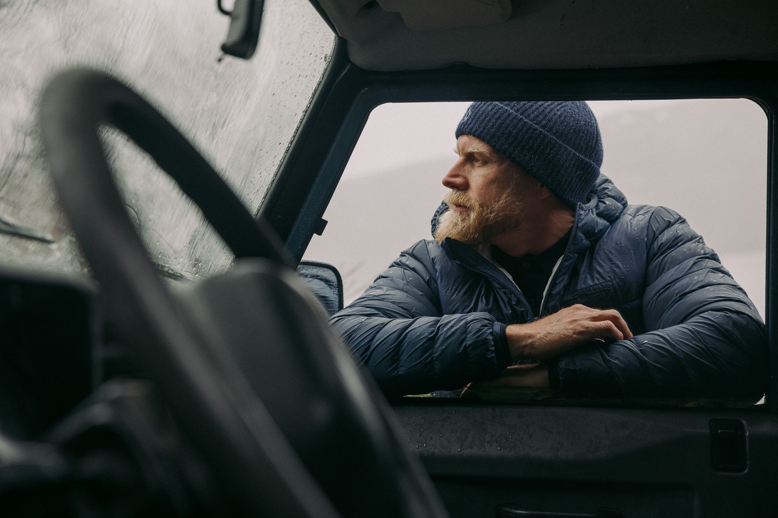 A man with a beard and a blue hat is sitting in the driver's seat of a vehicle, looking out the window at a snowy landscape.