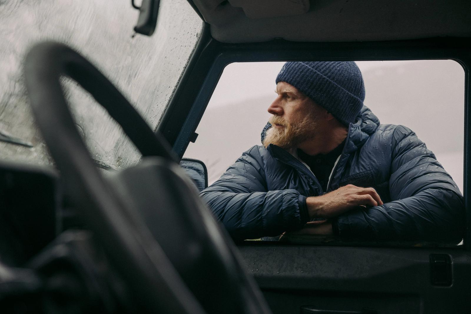 A man with a beard and a blue hat is sitting in the driver's seat of a vehicle, looking out the window at a snowy landscape.