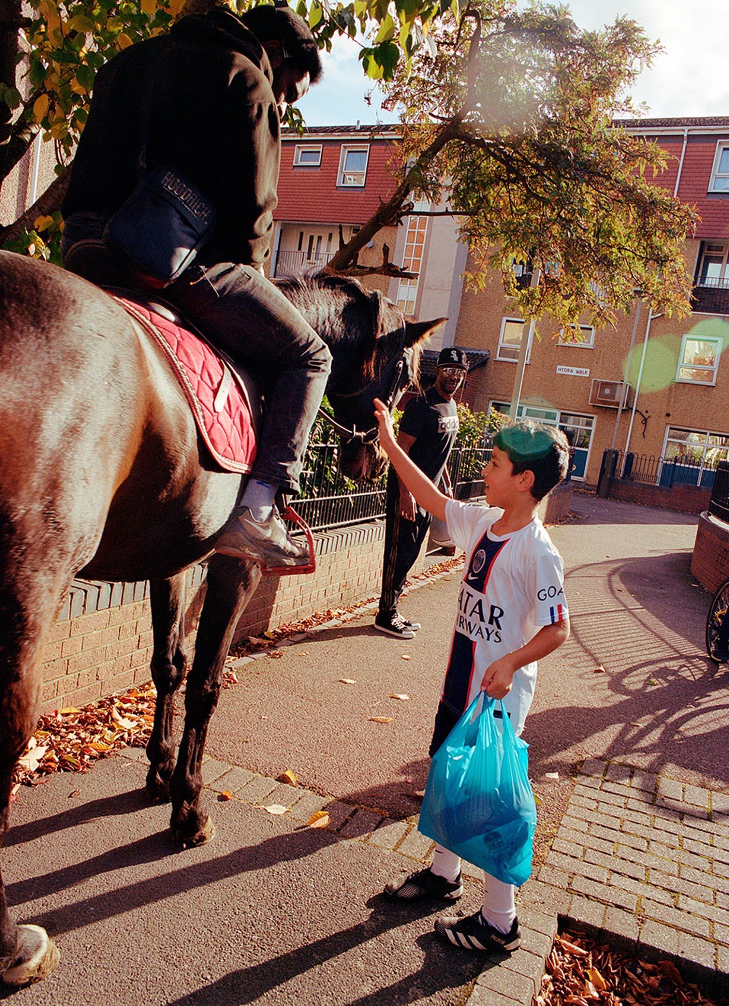 A young boy in a football jersey is reaching out to pet a horse in a residential street.