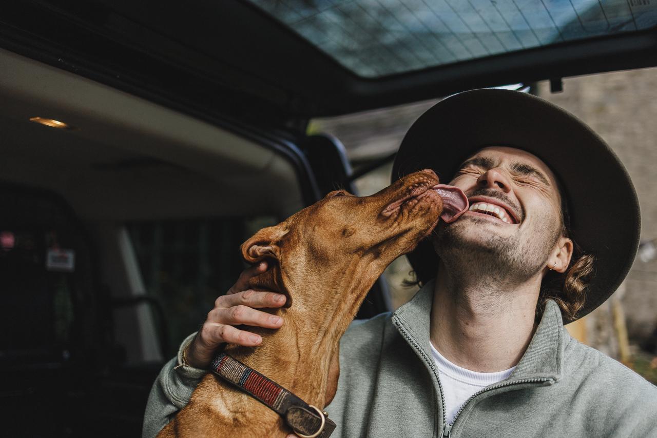 A man in a green jacket and hat is being licked by a brown dog.