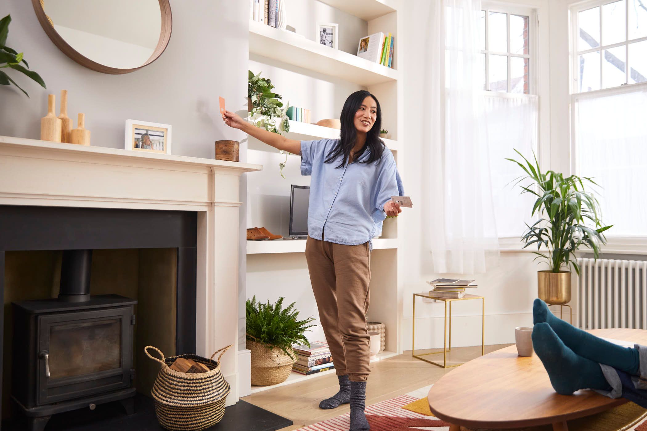 A woman with long dark hair is standing in a living room, holding a remote control and smiling at the camera. She is wearing a light blue shirt and brown pants.