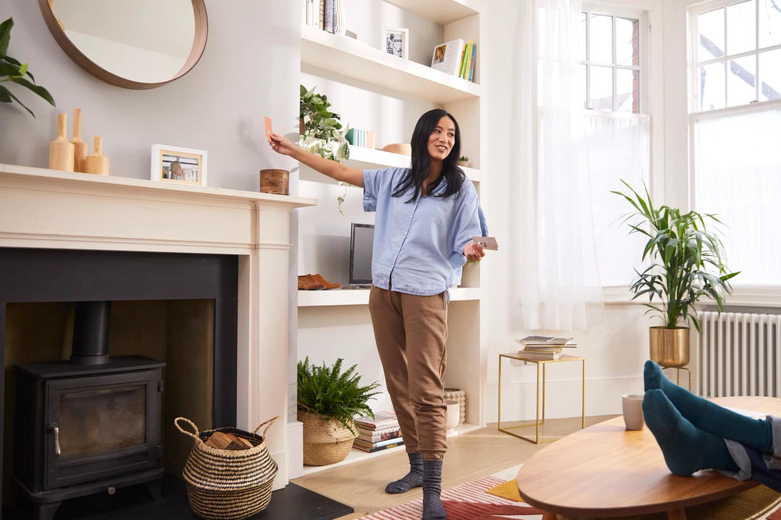 A woman with long dark hair is standing in a living room, holding a remote control and smiling at the camera. She is wearing a light blue shirt and brown pants.