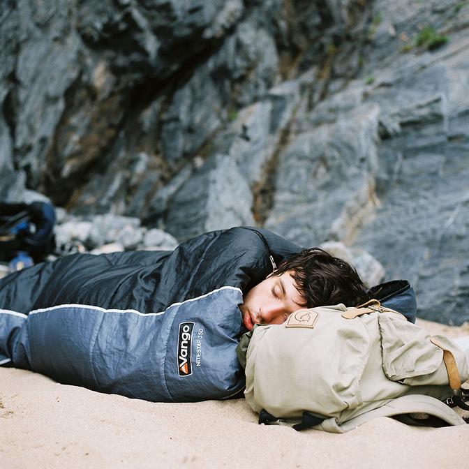 A person is sleeping on a beach, wearing a sleeping bag and a backpack.