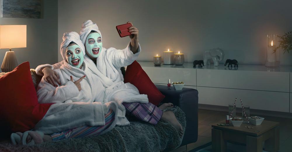 Two women in bathrobes take a selfie on a couch with candles and snacks nearby.