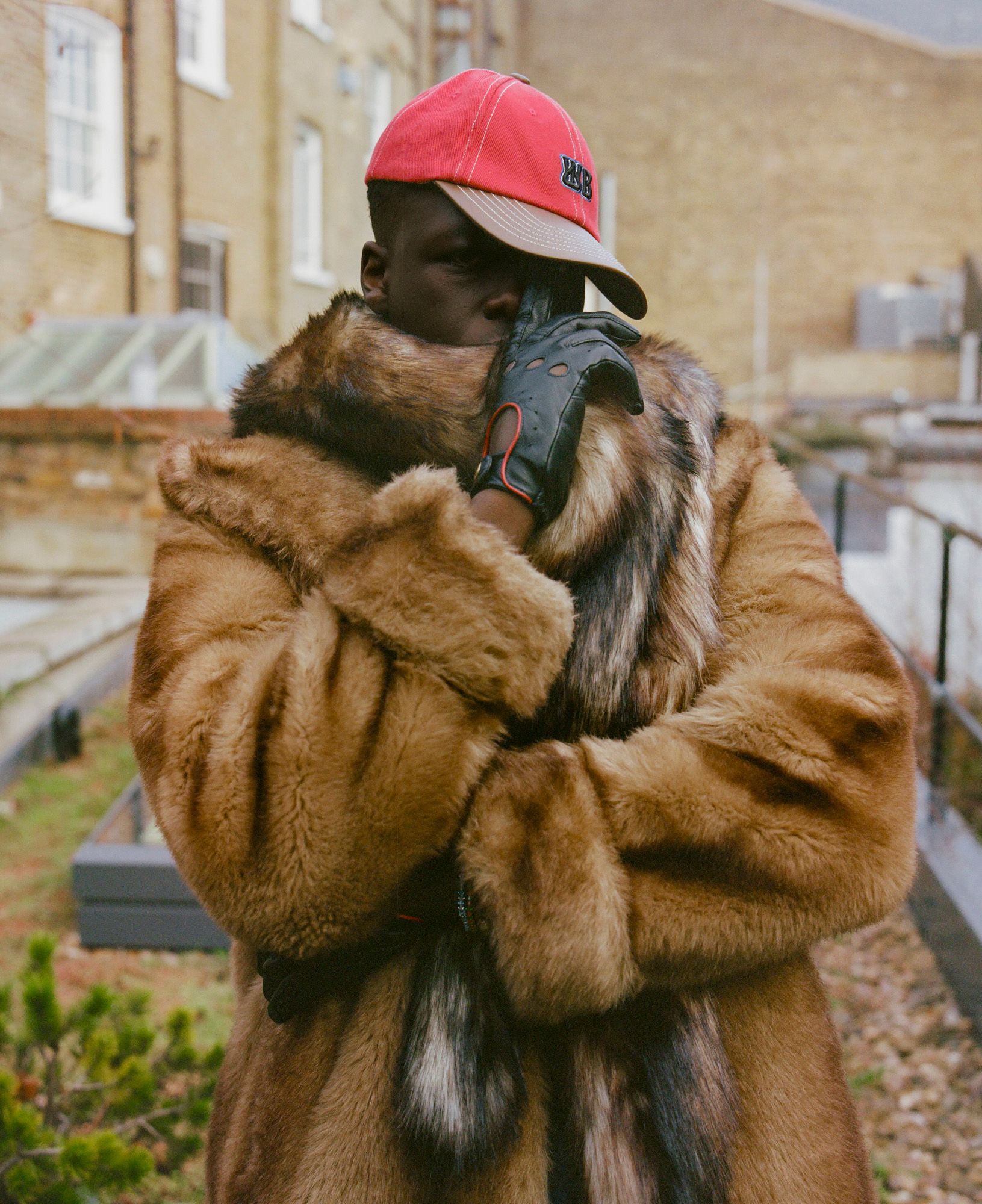 A person wearing a fur coat, a red baseball cap, and black gloves, standing in front of a brick building with a garden in the background.