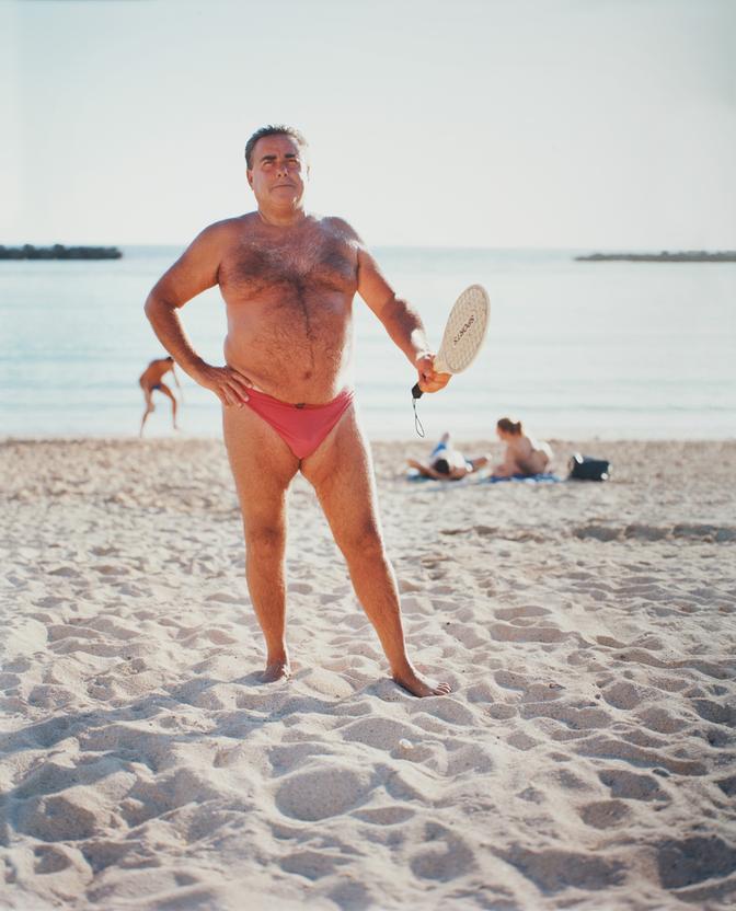 A man in a red swimsuit, also known as a 'speedo' or 'bathing briefs', is standing on a beach, holding a beach volleyball paddle.