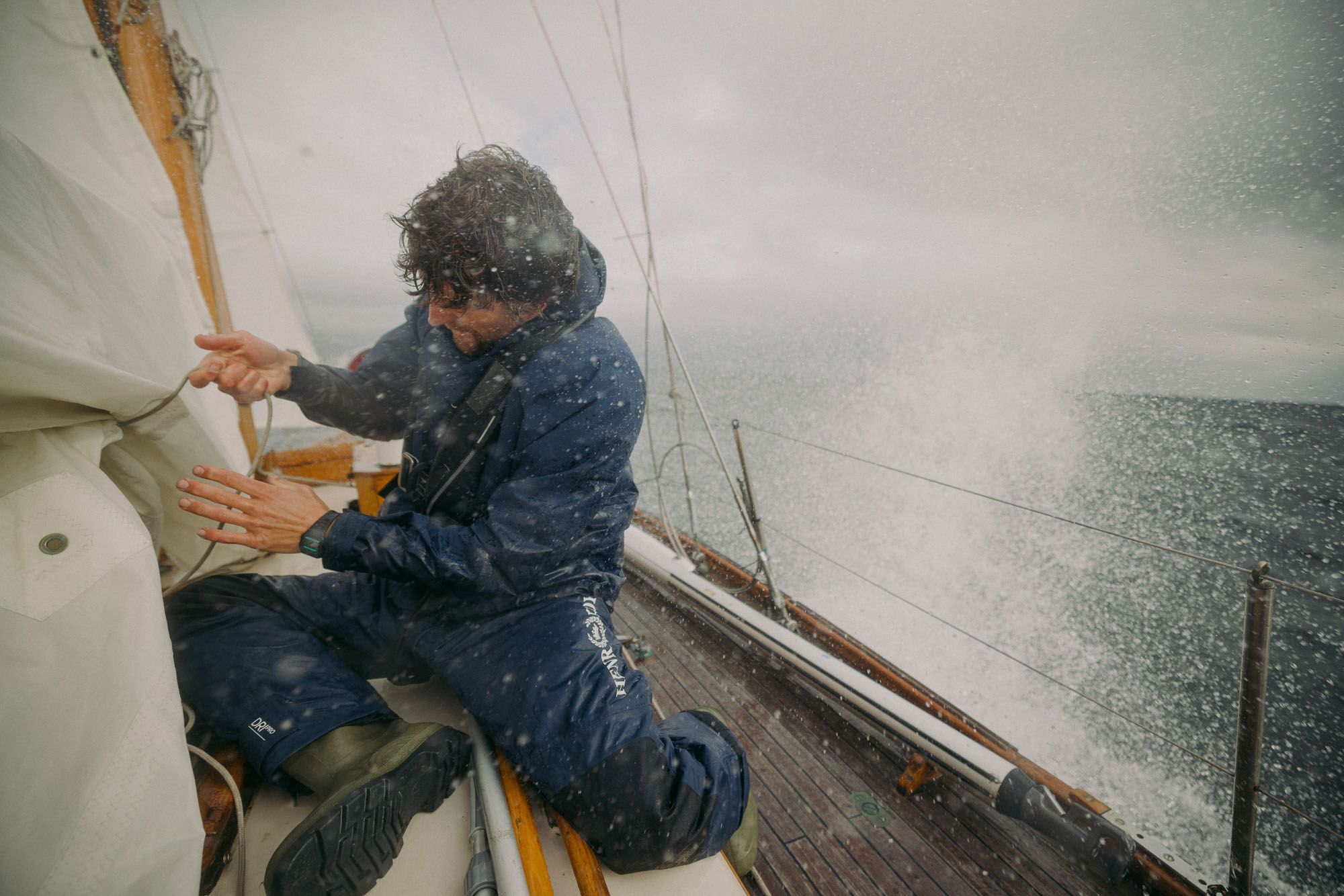 A man in a wetsuit and sailing jacket is sitting on a sailboat, holding onto the sail as the boat navigates through rough, stormy seas.