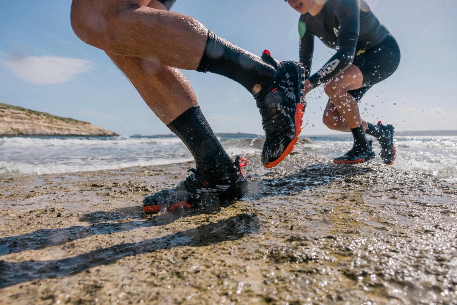 Two people in wetsuits running on a rocky beach, with the ocean in the background.