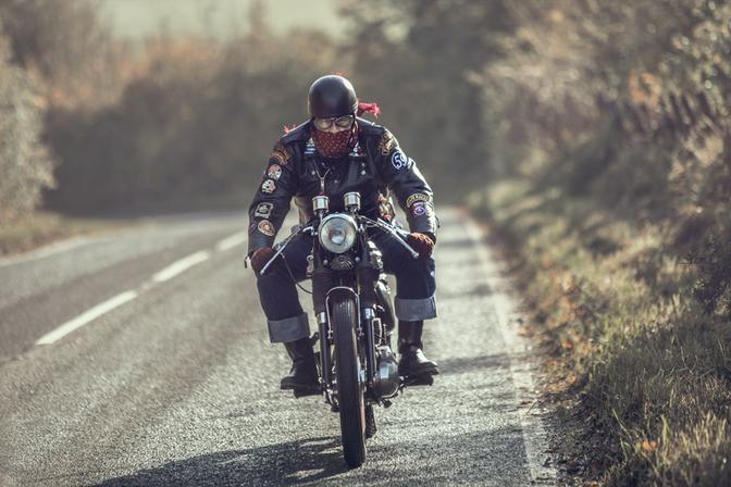 A biker wearing a black leather jacket with various patches, riding a motorcycle on a rural road.