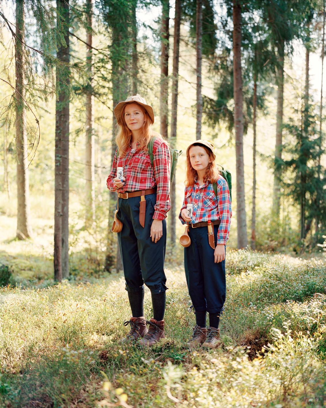 A woman and a young girl, both with long red hair, standing in a forest, both wearing plaid shirts, jeans, and hiking boots, holding small wooden cups.