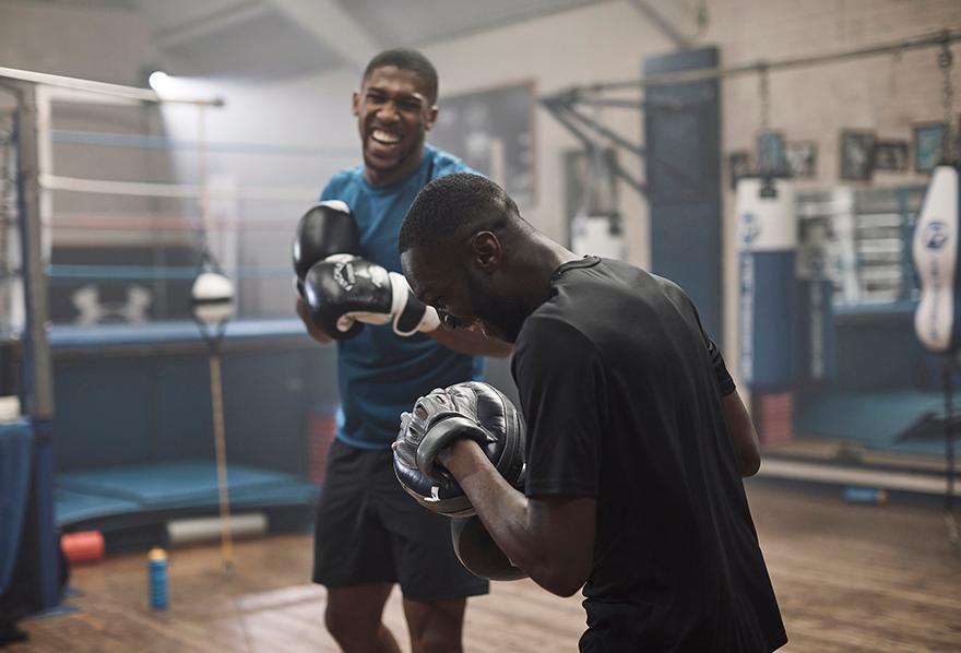 Two male boxers in a gym, one in a blue shirt and black shorts, the other in a black shirt and black shorts, both wearing boxing gloves and standing in a boxing ring.