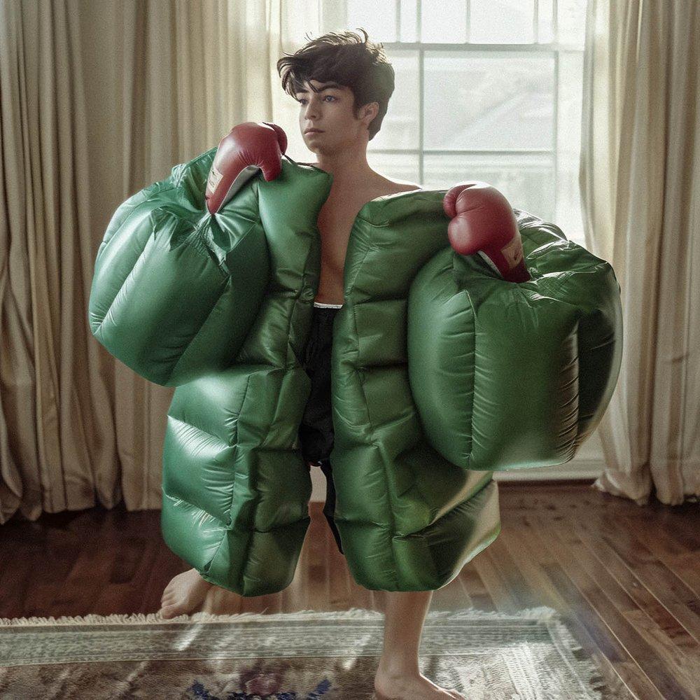 A young man wearing a green inflatable suit with boxing gloves, standing in a living room with a window in the background.