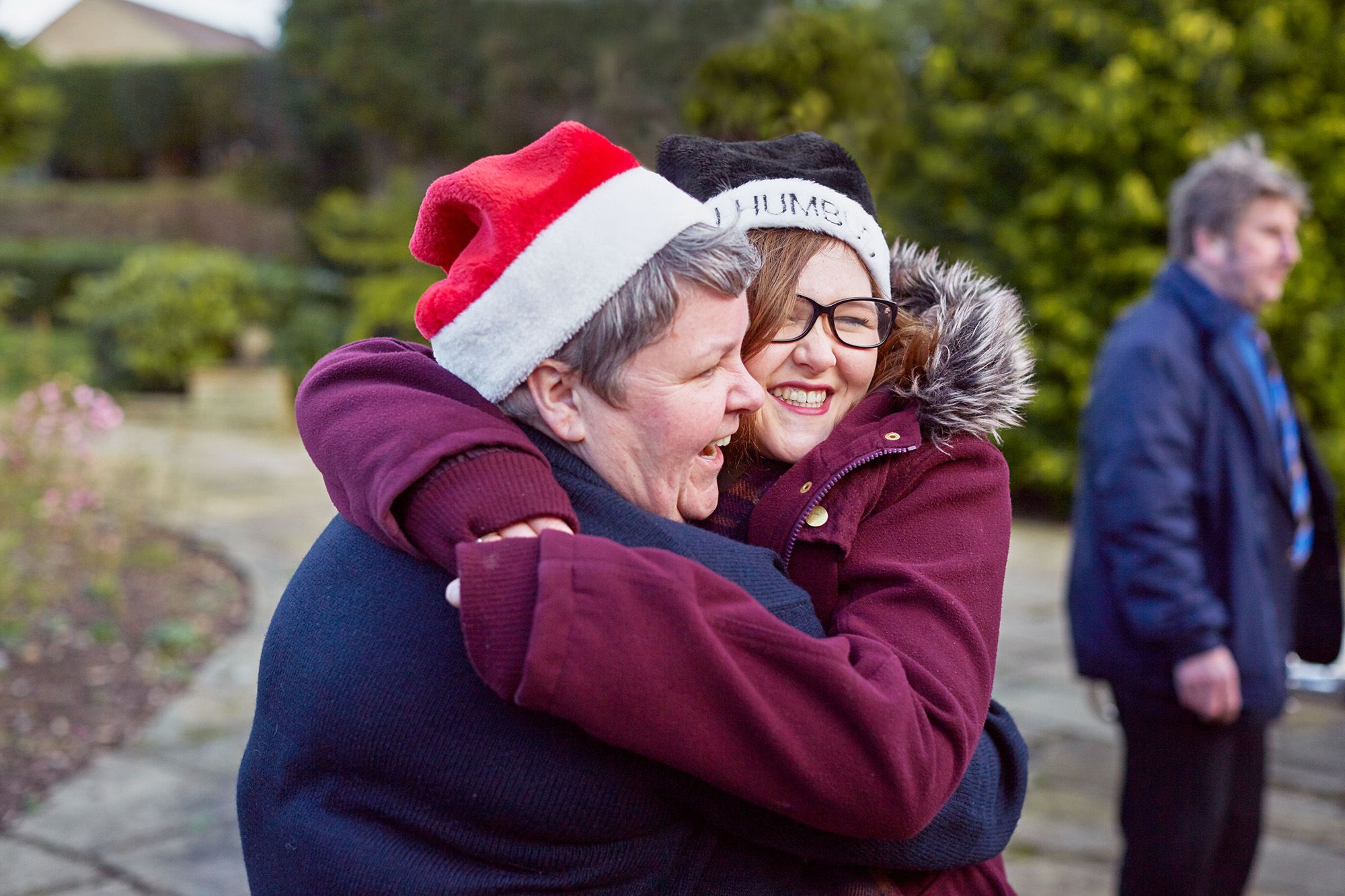 Two people embrace in a garden, both wearing Santa hats and winter coats.