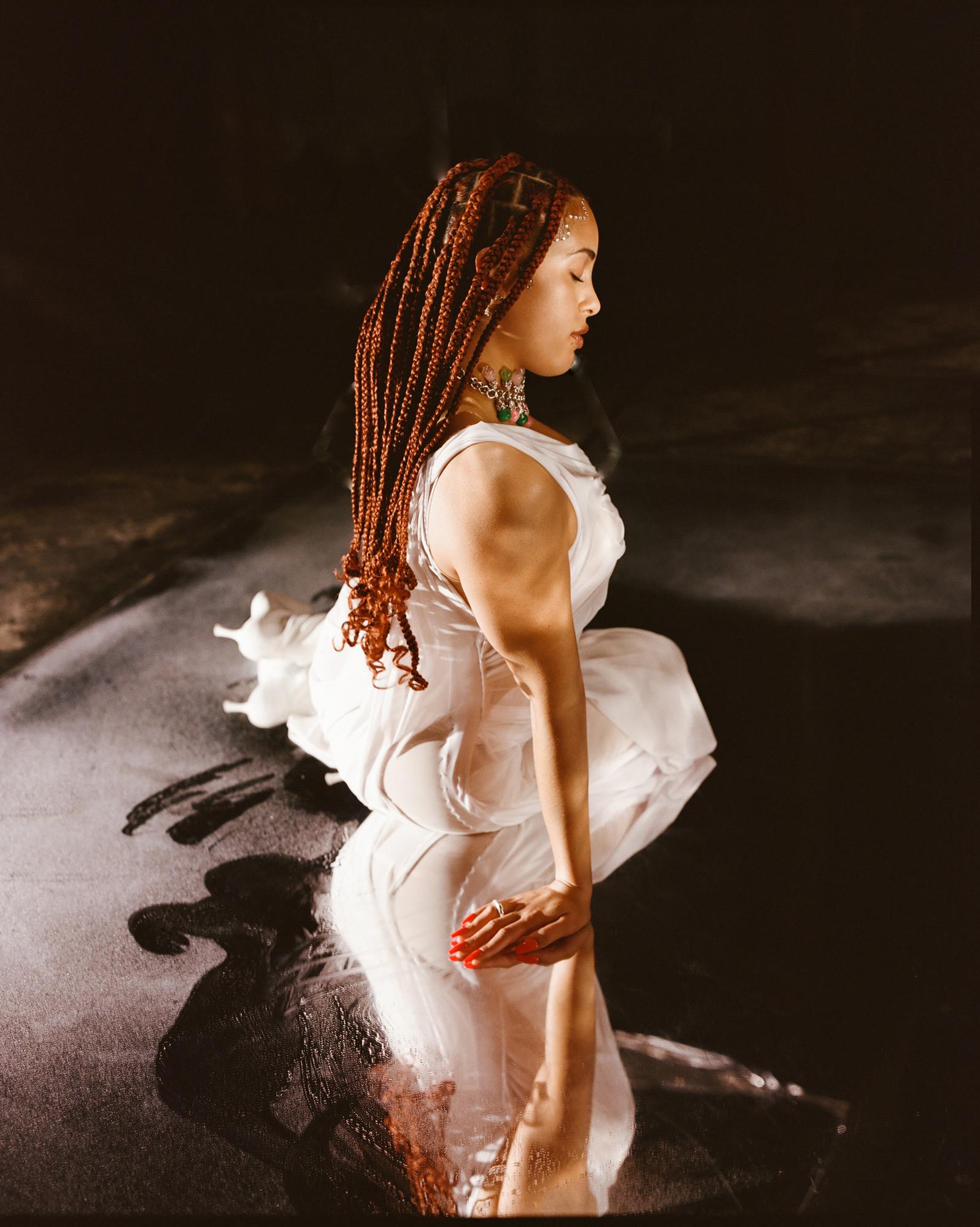 A woman with long braids and a white dress kneeling in a dark room, with her hands on her knees and her head down.