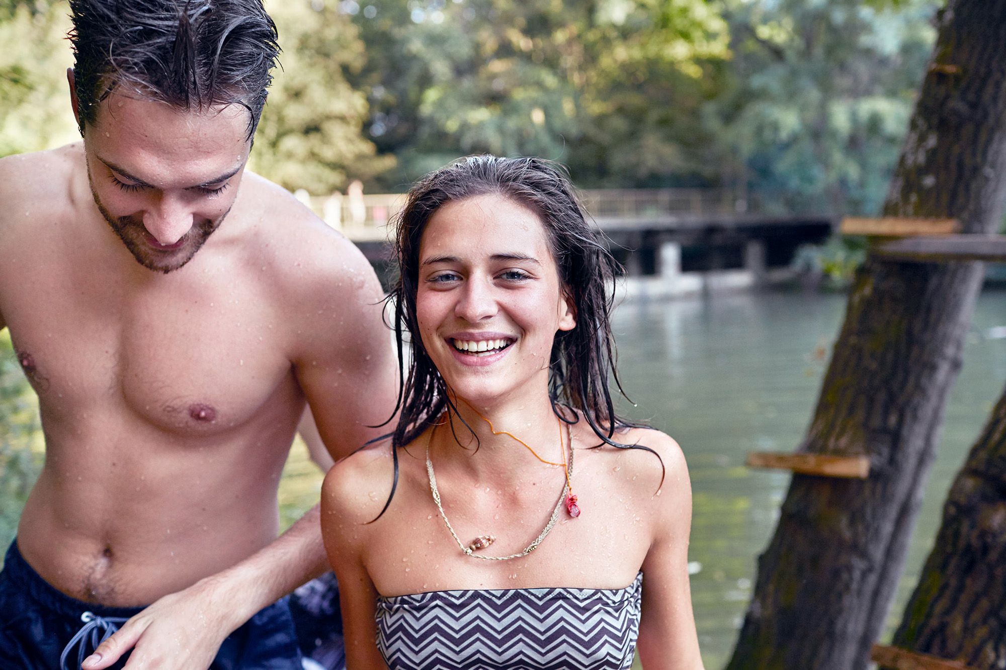 A woman and man standing near water, the woman smiles while the man stands behind her.