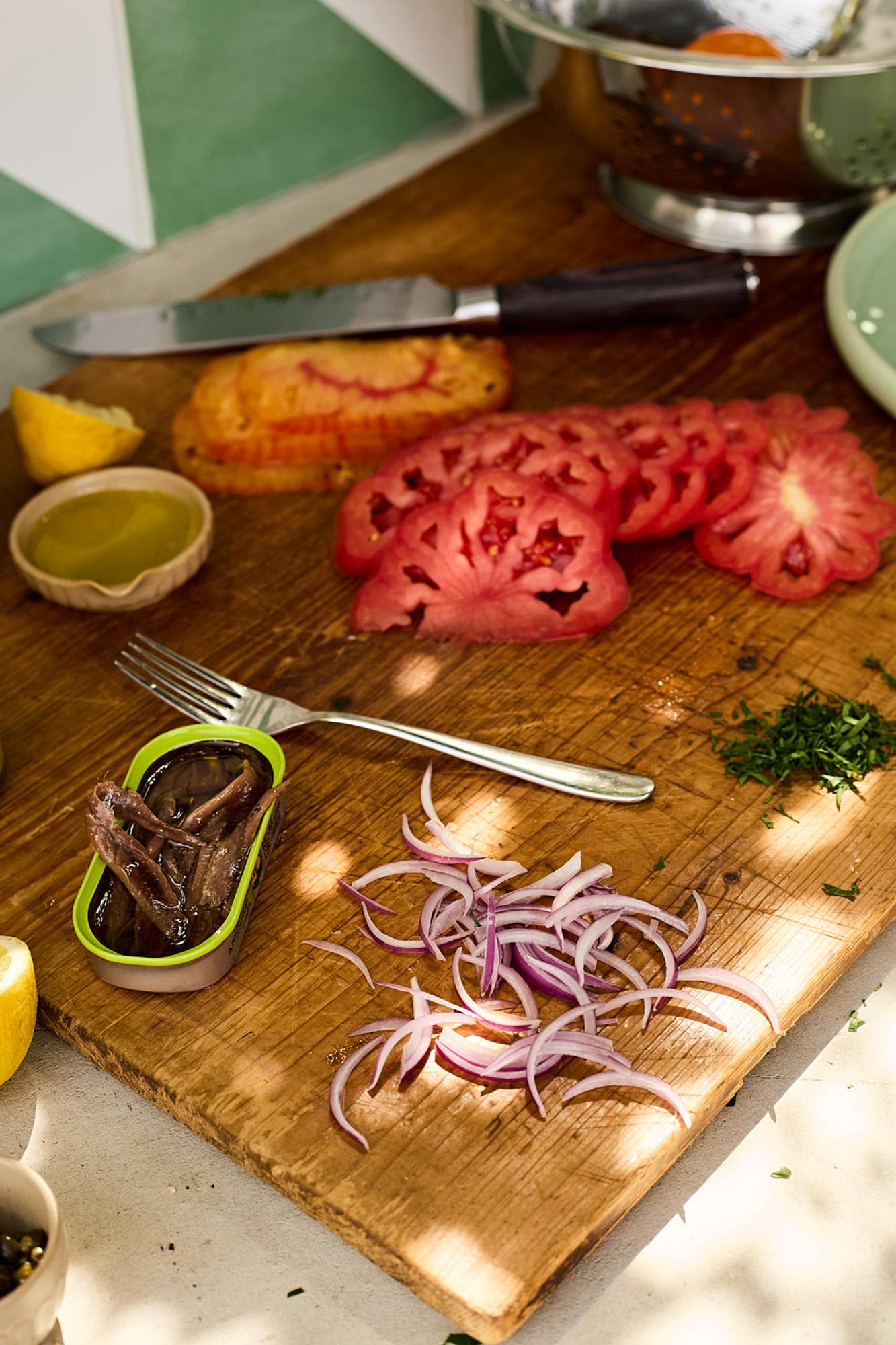 A wooden cutting board with sliced tomatoes, a knife, and a small bowl of olive oil, surrounded by various ingredients and cooking tools.