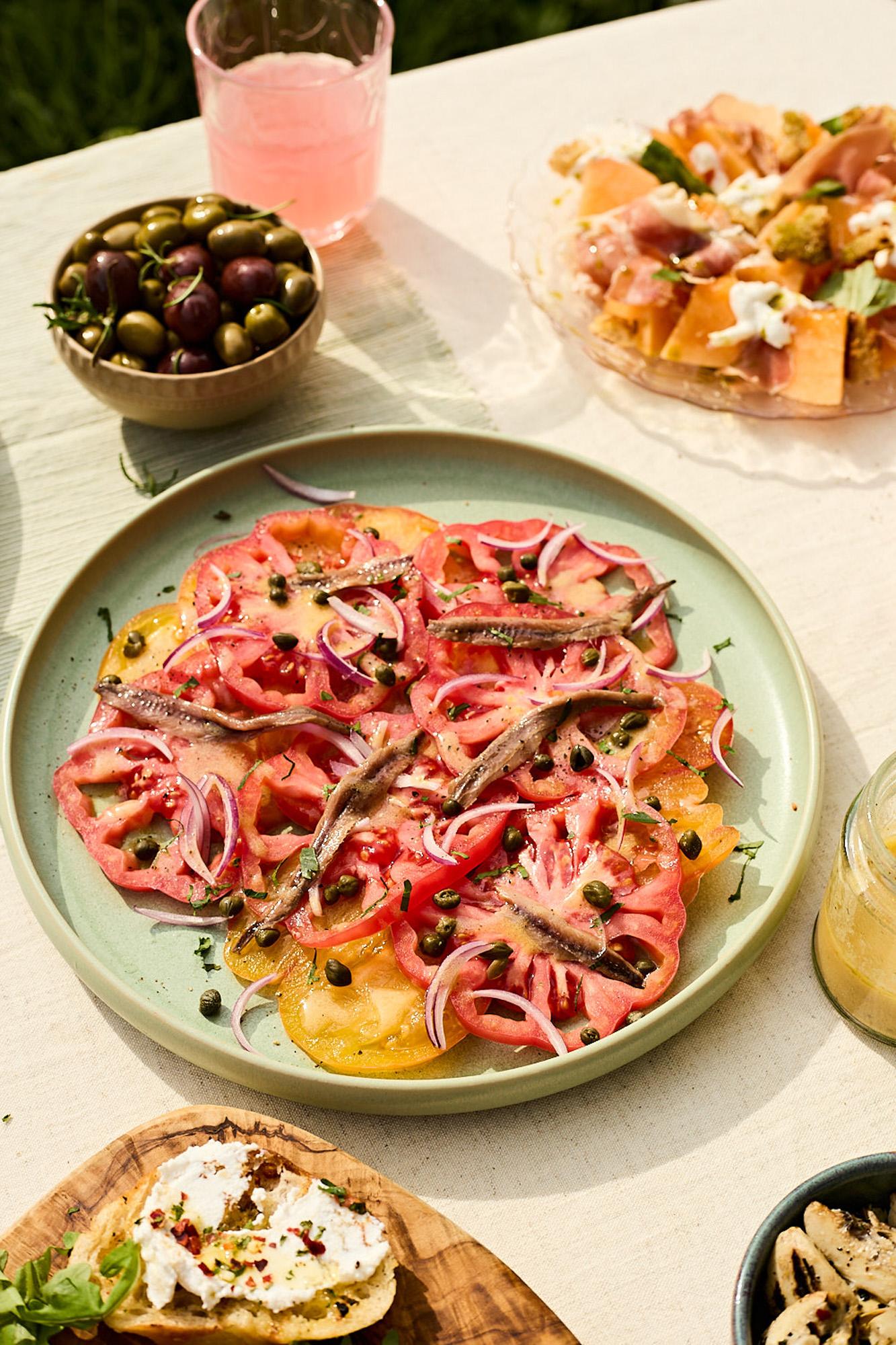 A plate of sliced tomatoes, capers, and anchovies, garnished with fresh herbs, on a white tablecloth in a sunny outdoor setting.