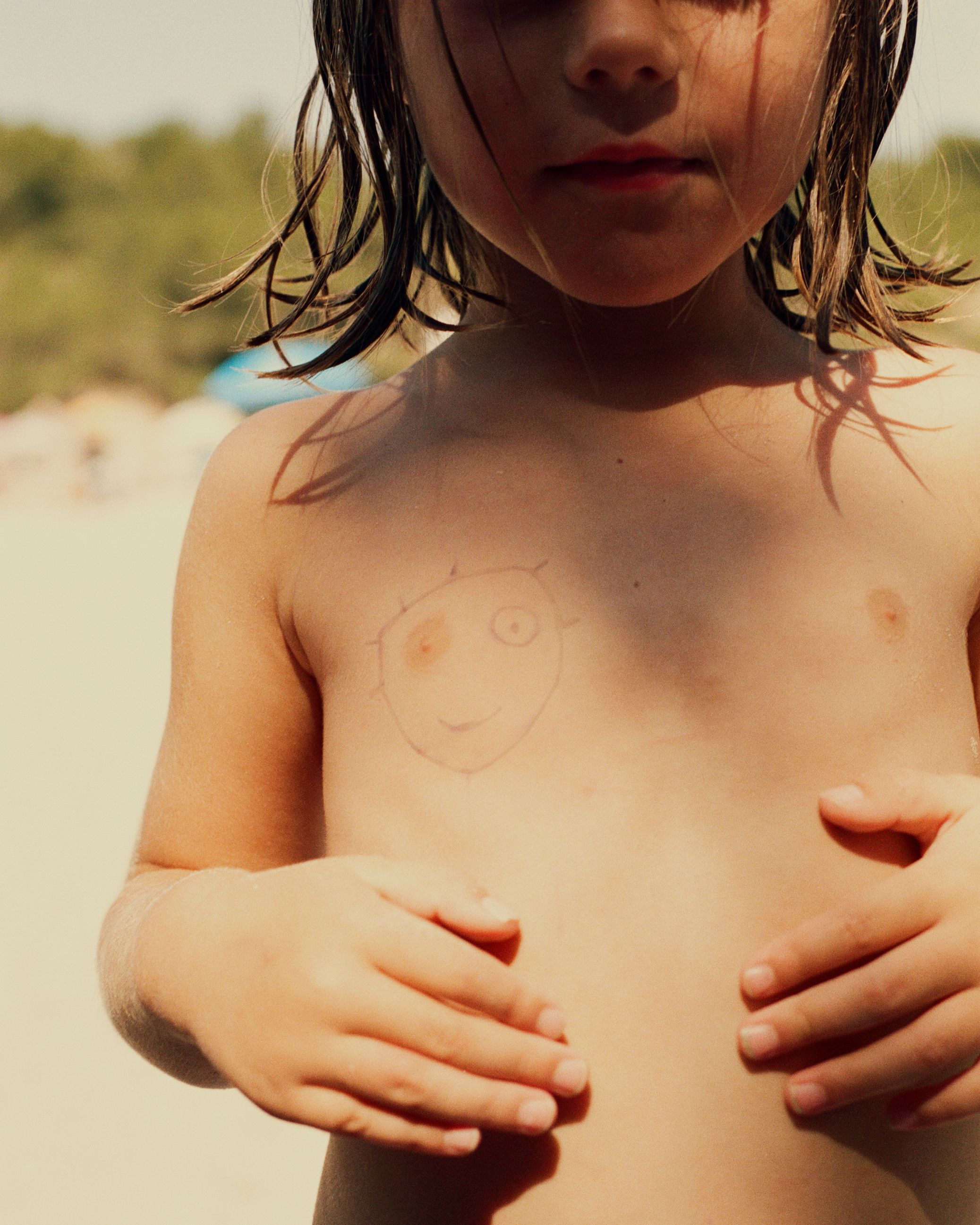 A young girl with a crude, hand-drawn, cartoon-style face on her chest, likely in a beach or pool setting, wearing a swimsuit or beachwear.