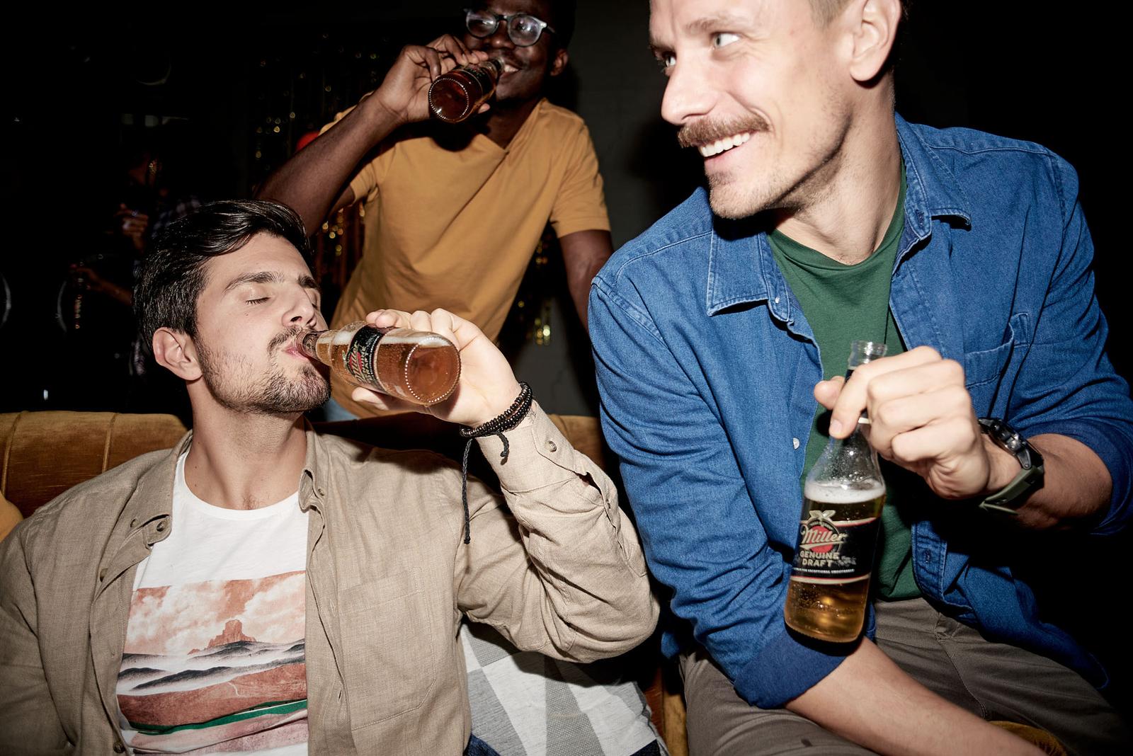 Three men in casual clothing, two holding beer bottles, one smiling and one looking at the camera, in a dimly lit background.