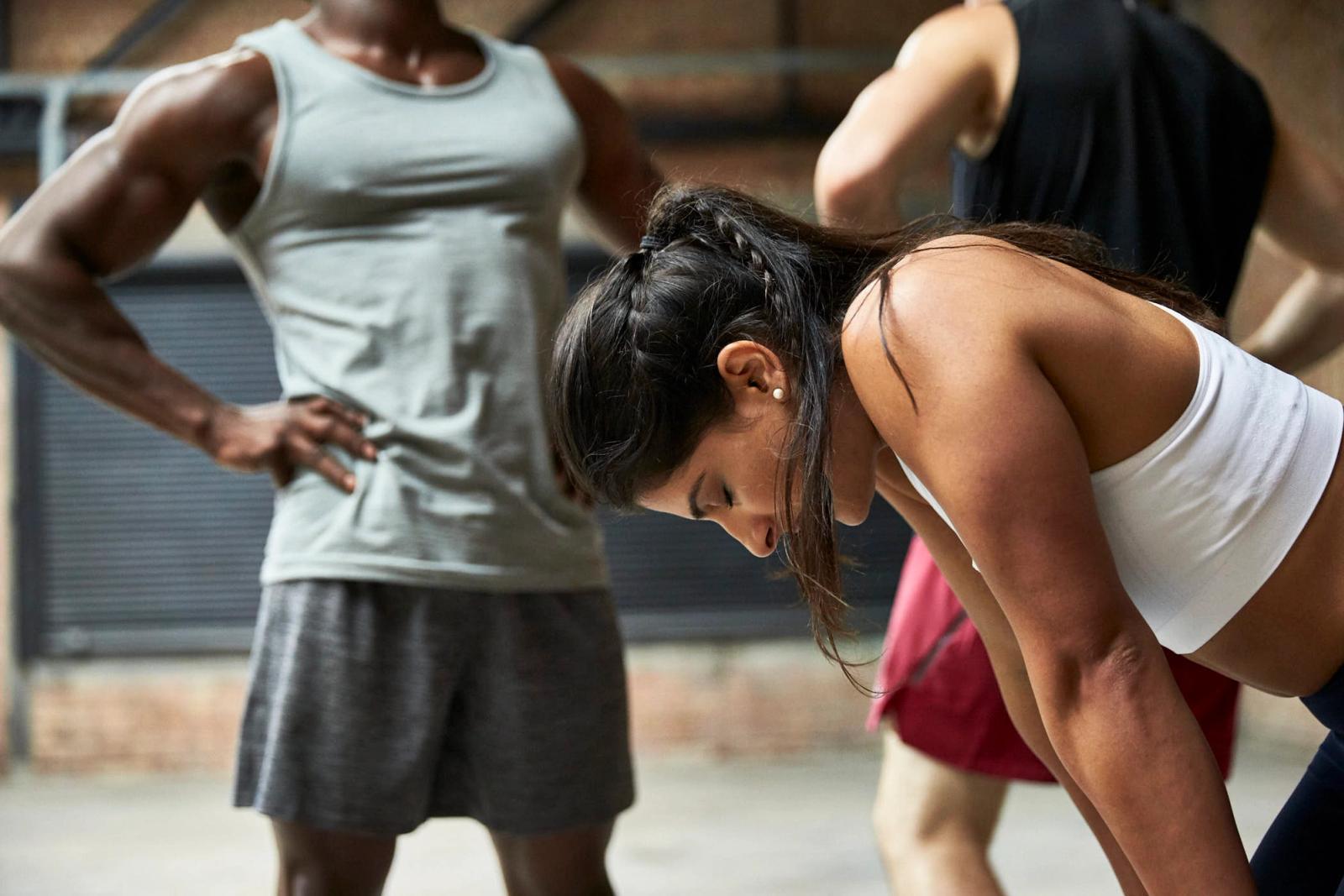The woman in the image is bent over, gasping for breath, her face red from exertion. She is surrounded by others who appear to be exercising or working out in a gym or fitness.