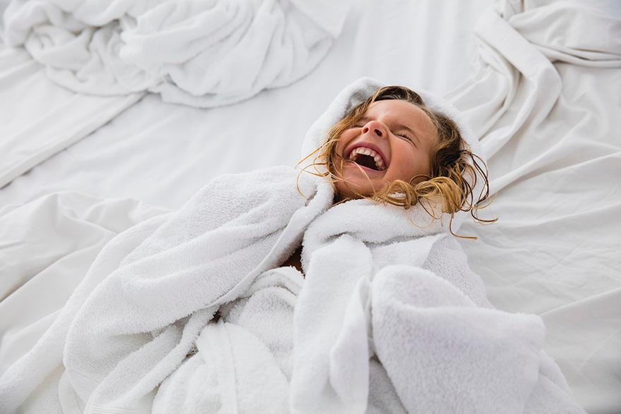 A young girl in a white bathrobe, laughing and lying on a bed with white sheets.