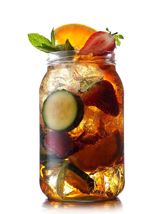 A glass of iced tea with a slice of orange, a sprig of mint, and a few ice cubes, with a glass of iced tea in the background.