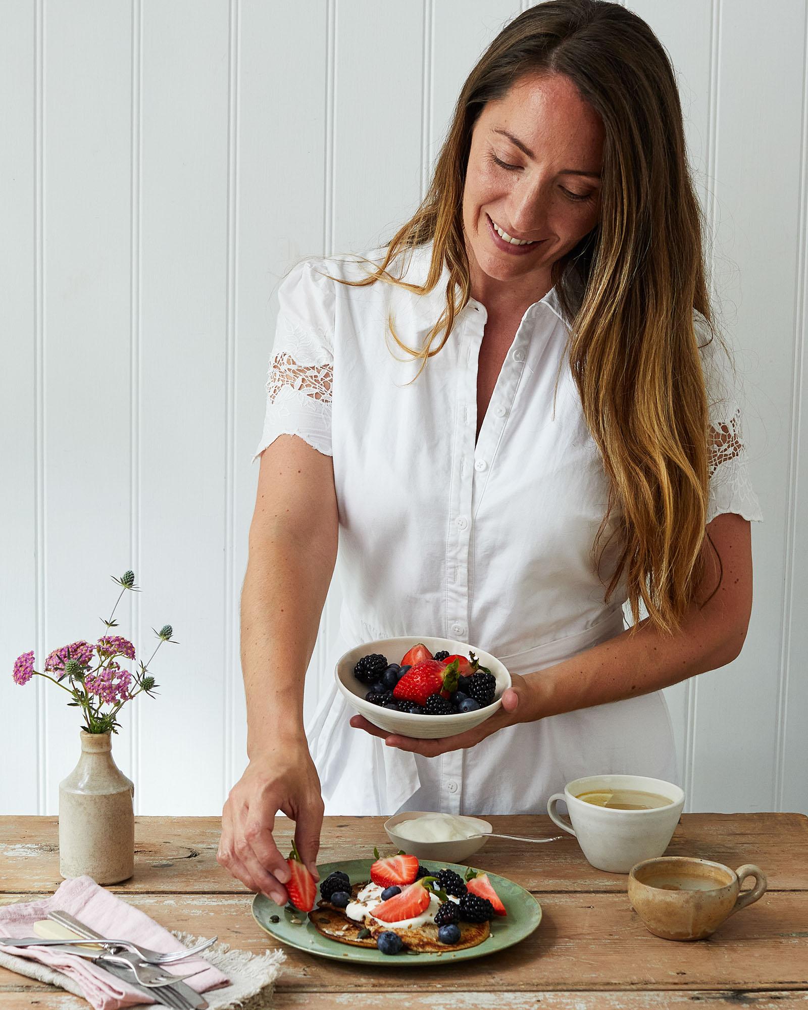 A woman places a strawberry on a plate of pancakes topped with fruits and whipped cream.