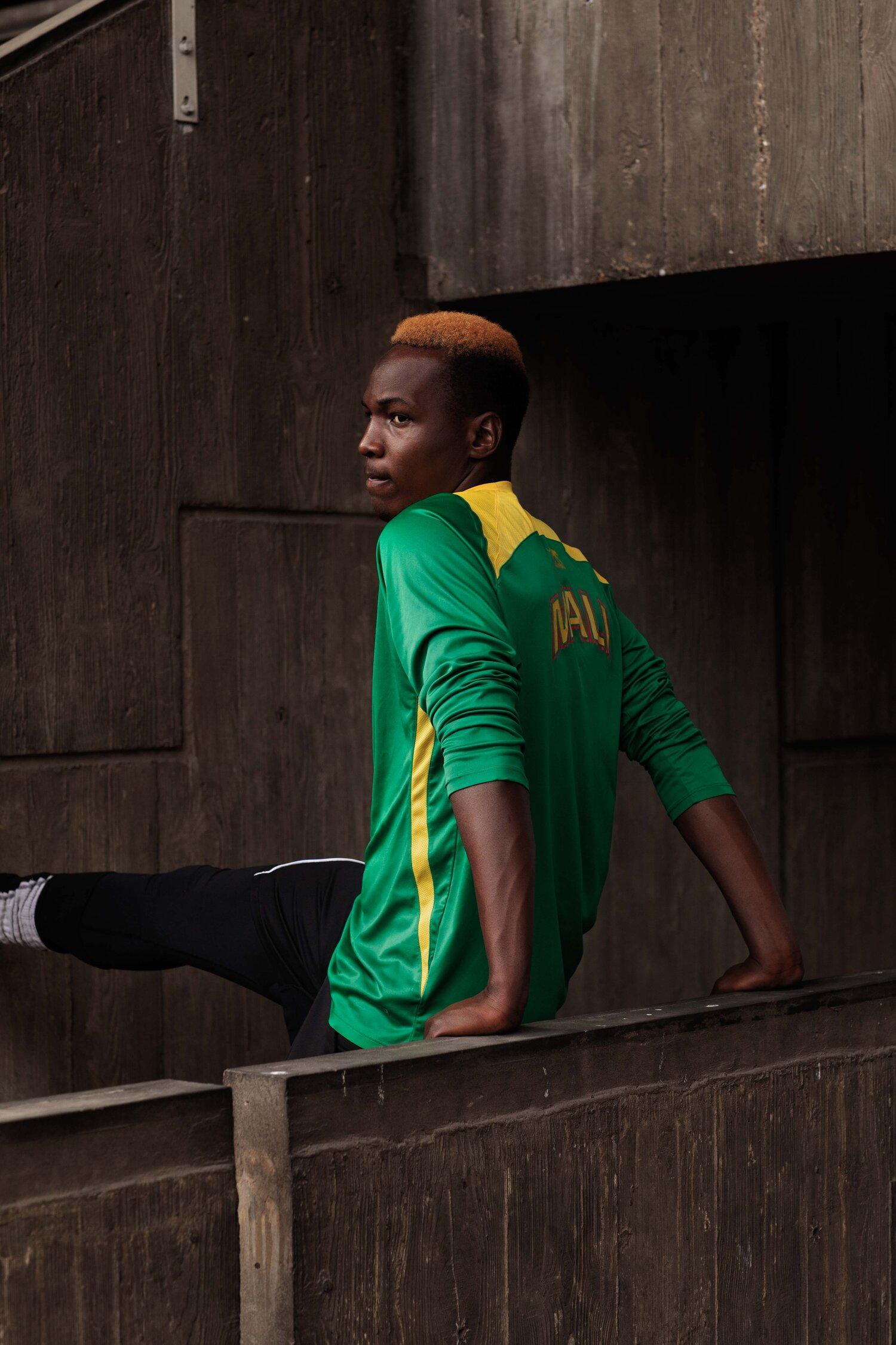 A young man with short, spiky hair stands in a dark industrial setting wearing a green and yellow jersey with 'MAD' on the back.