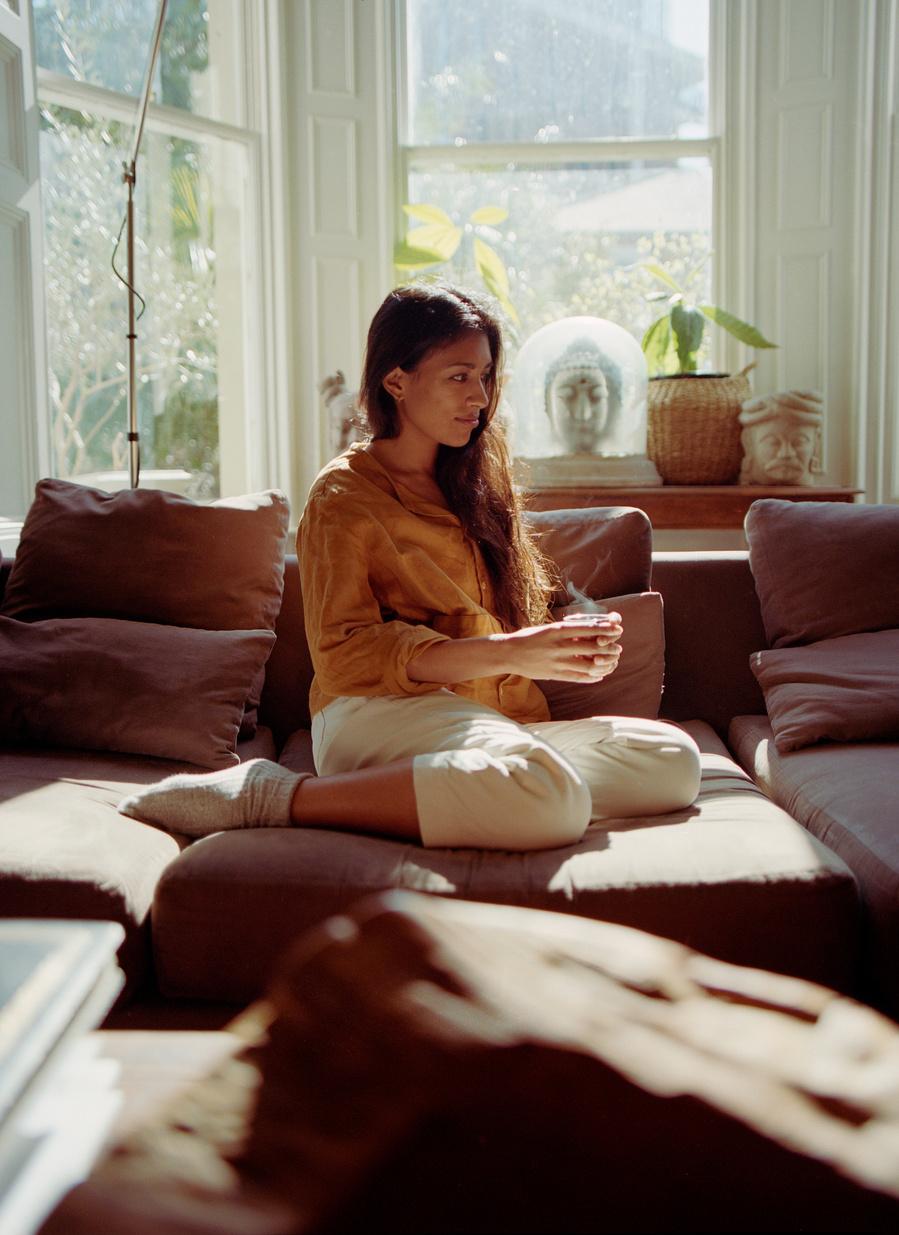 A woman sitting on a couch, wearing a yellow blouse and white pants, holding a cup of coffee in a sunny living room.