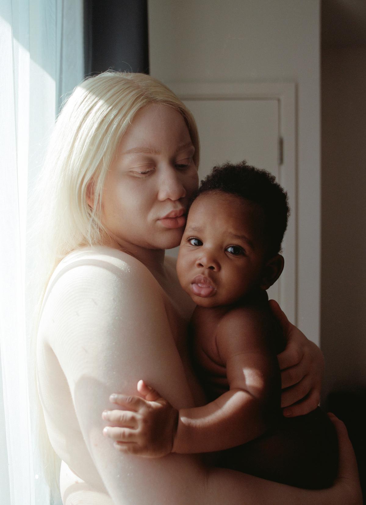 A woman with blonde hair holding a baby with dark skin, both naked, in a dimly lit room with a white door in the background.