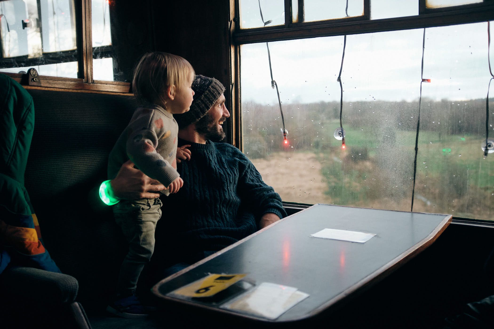 A man and a child sit together on a train, gazing out the window at a rural landscape. The man, wearing a dark green sweater, has his arm around the child, who is dressed in a.