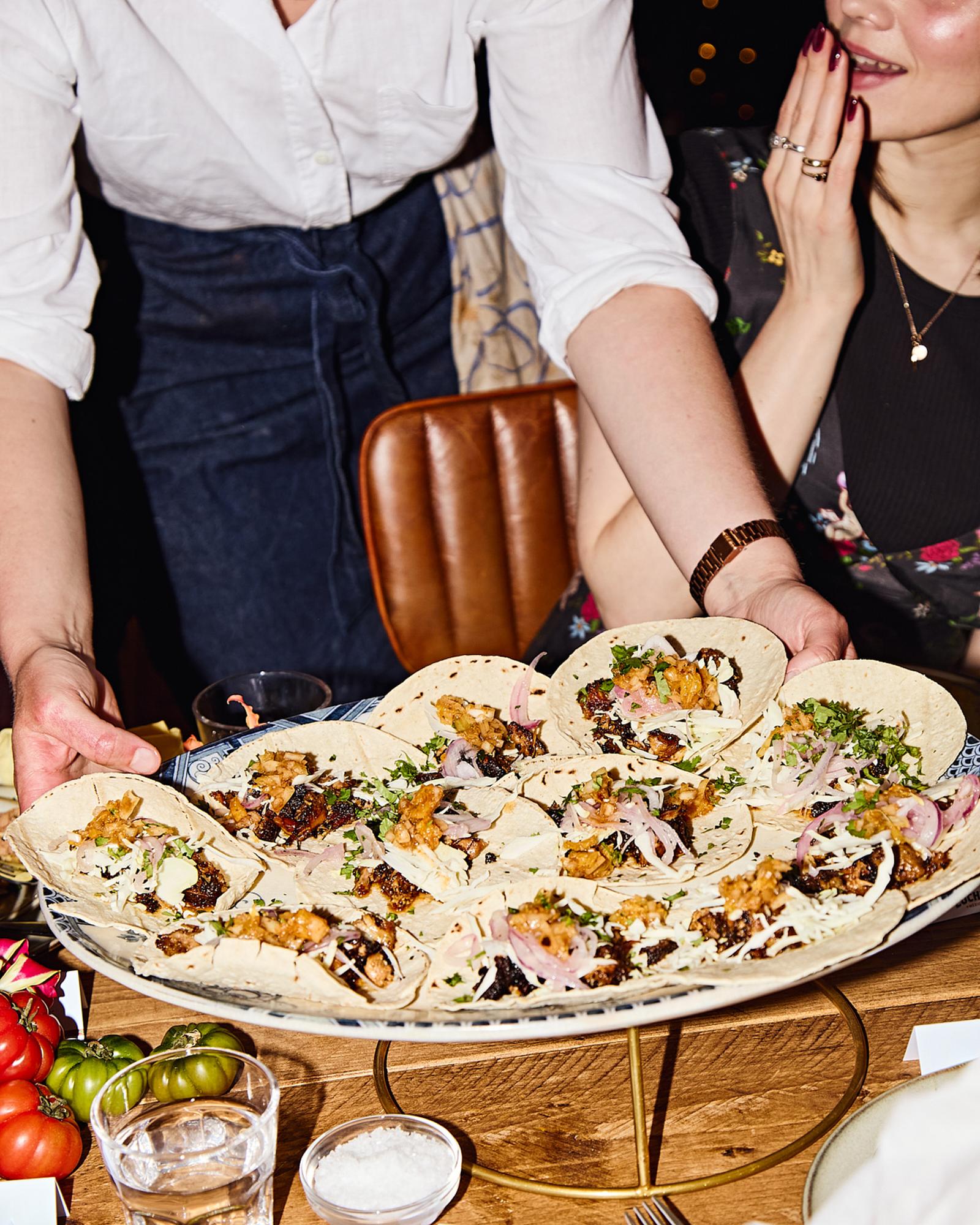 A woman in a white shirt and dark jeans is holding a large platter of tacos, with a few other food items and a glass of water in the background.