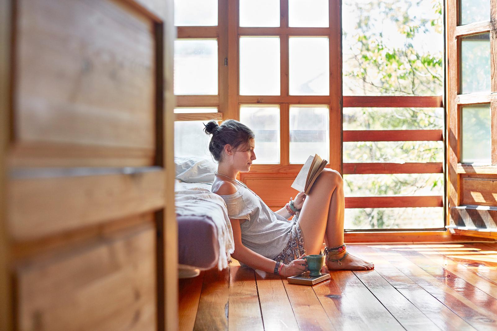 A woman sits on the floor, reading a book and holding a cup of coffee, in a cozy room with a large window and a comfortable-looking bed.