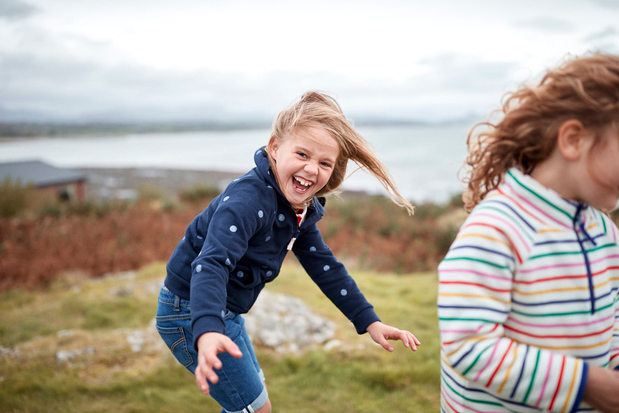 A young girl with blonde hair and a big smile, wearing a navy blue sweatshirt with white polka dots and blue jeans, is running towards the camera with her arms outstretched and.