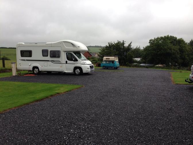 A parking lot with several vehicles, including a white motorhome, a blue camper van, and a few cars, in a rural or countryside setting.