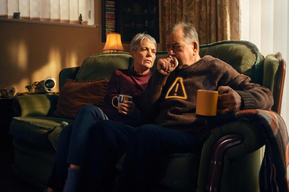 An elderly couple sitting on a couch, both holding cups and looking at something in the distance, likely a TV or a window with a view.