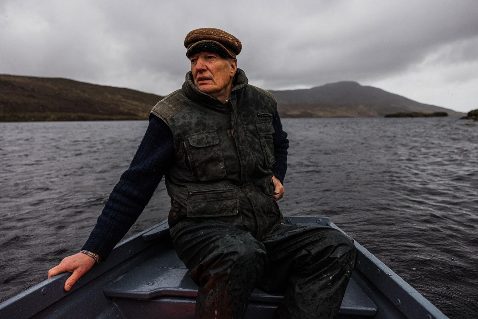 A man in a brown hat, black jacket, and dark trousers is sitting in a small, blue boat on a lake, with a mountain range in the background.